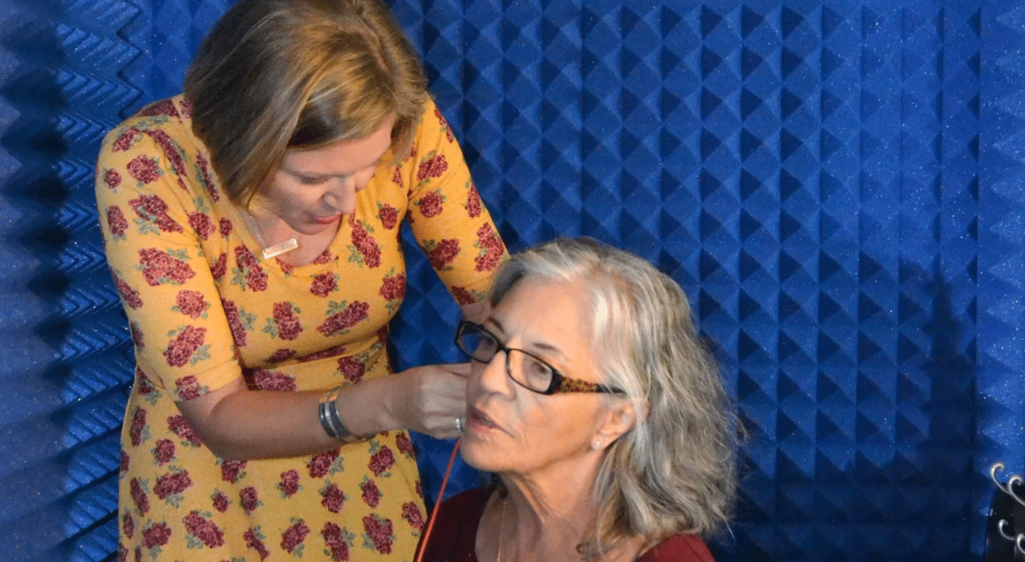 Audiologist conducting a hearing evaluation on an elderly woman inside a WhisperRoom audiometric booth, featuring blue acoustic foam on the interior walls.