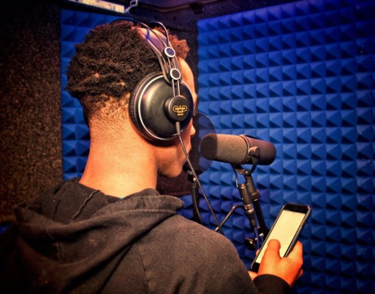 A young man wearing headphones and speaking into a microphone while recording inside of a WhisperRoom portable vocal booth