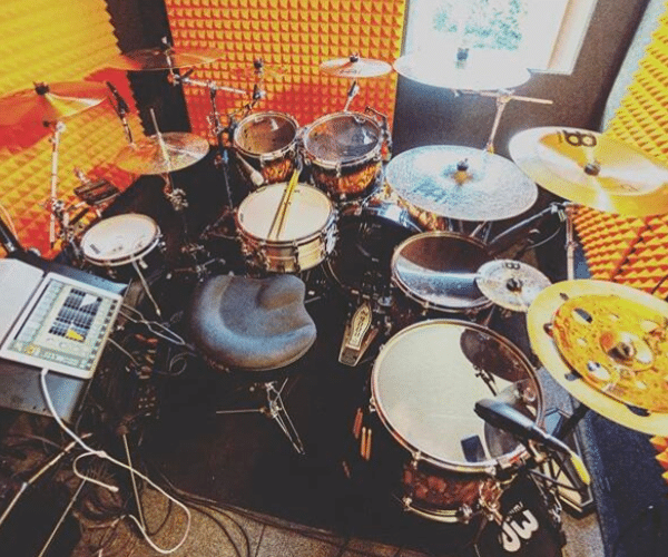 Image of a full drum kit and a laptop inside a medium-sized WhisperRoom drumming booth. The room features orange acoustic foam along the interior walls.