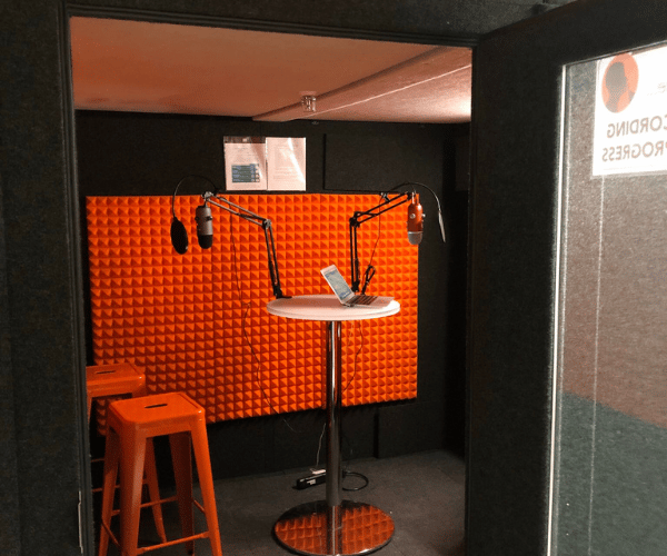 A WhisperRoom podcast booth shown with several microphones on a round table and a couple of stools.
