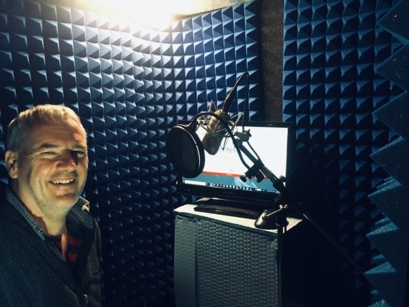 Professional voice actor Ian Russell smiling next to his microphone setup, inside of his home studio's portable vocal booth.