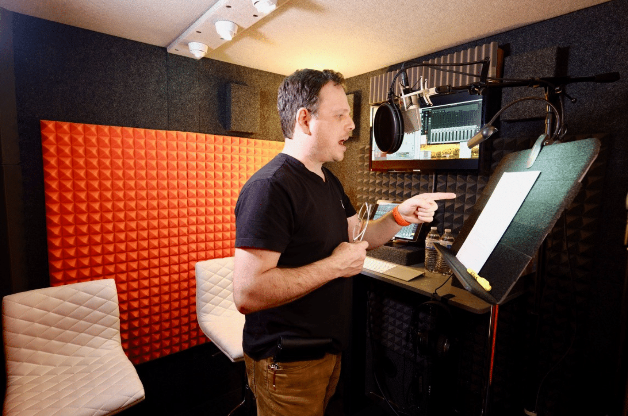 Man standing inside his WhisperRoom vocal booth, recording a voice-over take. A computer is mounted to the interior wall, and he reads a script placed on a music stand.