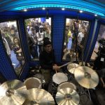 A crowd looks through windows to observe a drummer inside of Go-Pro's custom built WhisperRoom at the 2016 NAMM Show