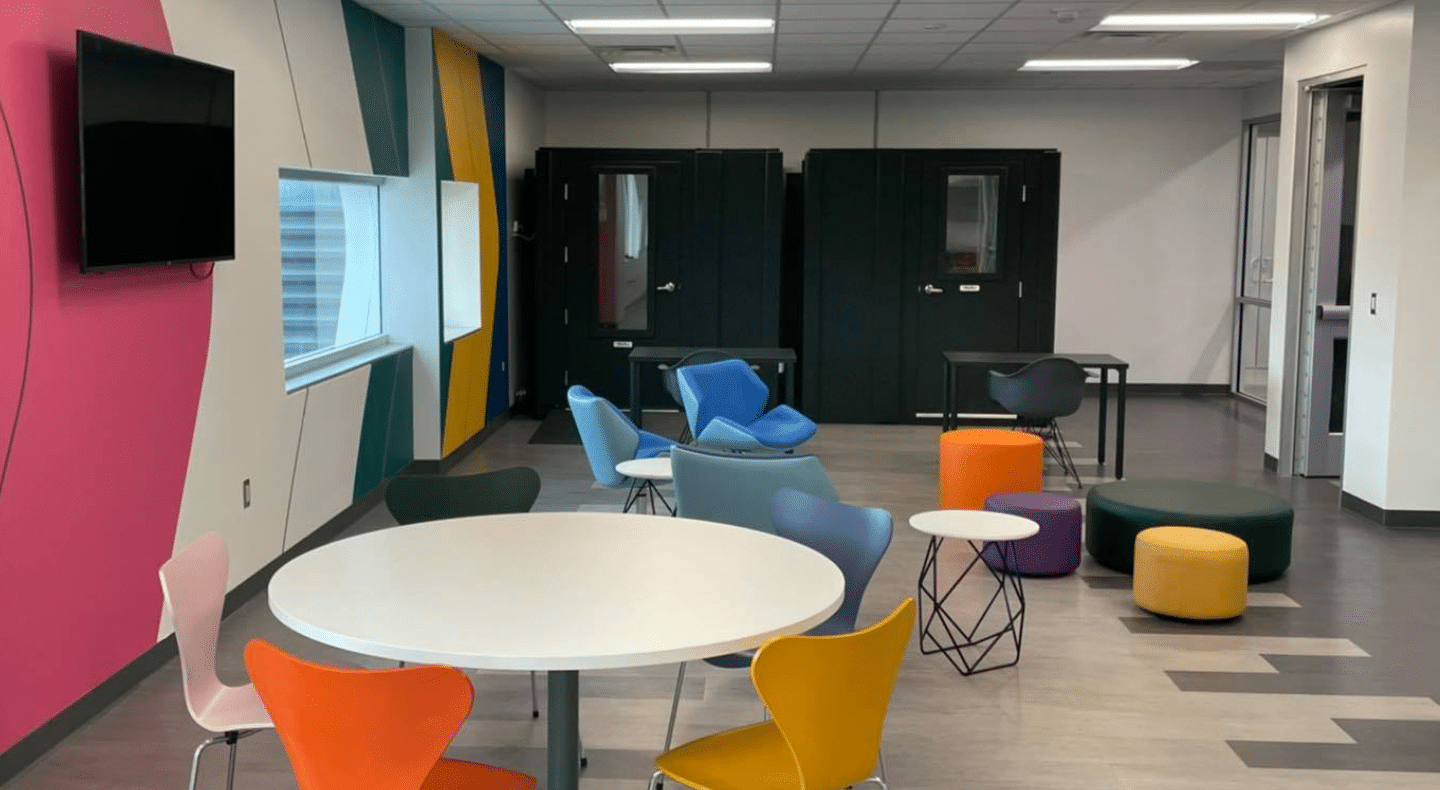 Two 6' x 6' WhisperRoom Privacy Booths in an open floor office space. Seating area with circular tables and multicolored chairs visible outside the booths.