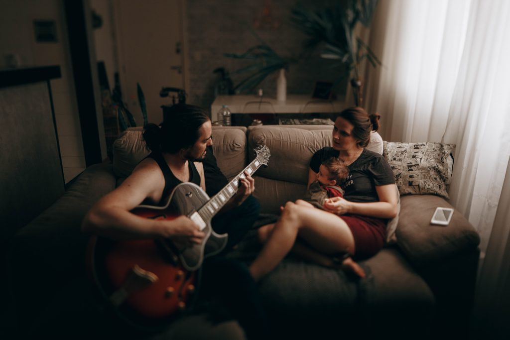 A man playing guitar on the couch with wife and baby sitting next to him