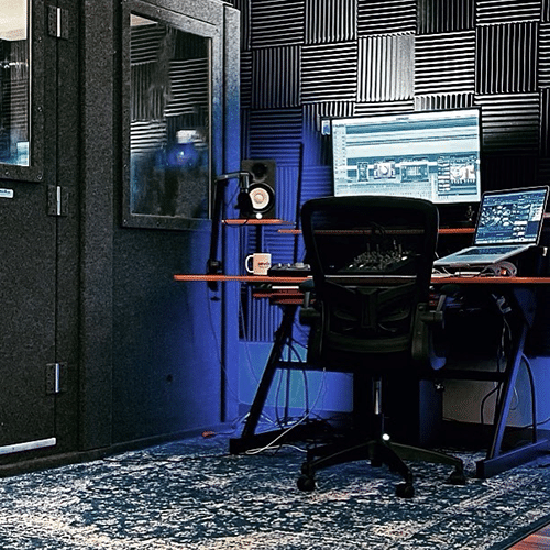 A recording desk with computers, speakers, a microphone, and chair set up in a studio next to a WhisperRoom.