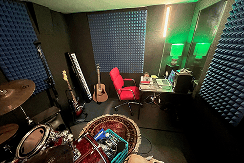 The interior of a WhisperRoom MDL 102126 S is shown with a desk that's set up for recording, a couple of guitars, a drum set, and more equipment.