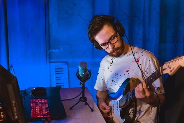 A man recording electric guitar at a desk in a small home recording studio.