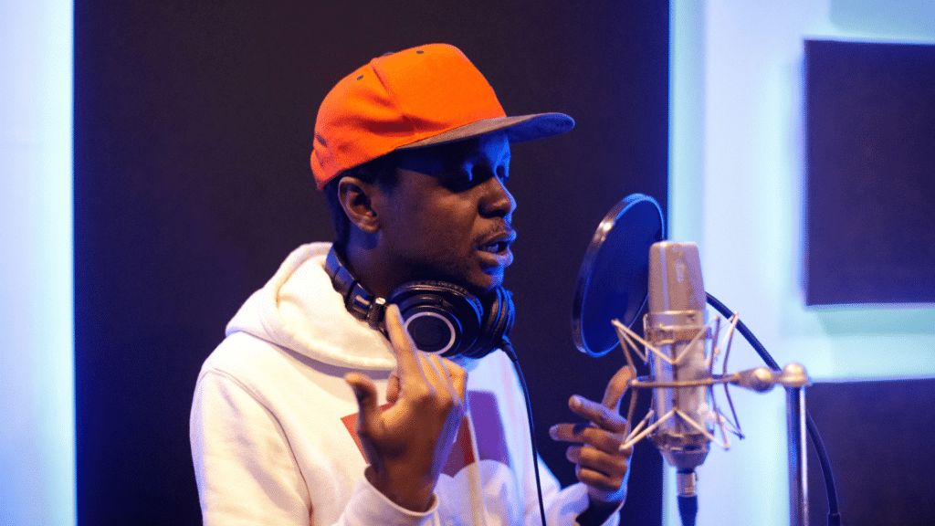 Image: A man recording vocals into a condenser microphone in a recording studio, wearing headphones around his neck and an orange ball cap.