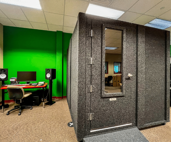 A single-wall WhisperRoom MDL 7296 S isolation sound booth, measuring 6' x 8', featured prominently in a high school's creative space. Adjacent to the booth is a well-equipped recording desk, complete with studio monitors and various recording gear.