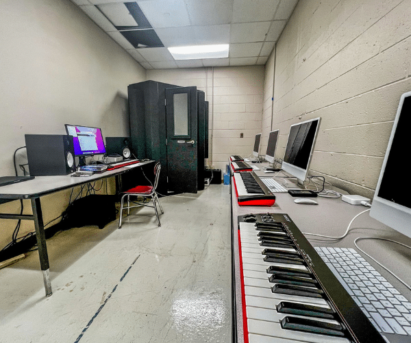 Image features a WhisperRoom MDL 127 LP S, a low-profile corner unit vocal booth, positioned inside a high school's audio production and creative skills classroom. A row of desktop computers and MIDI controllers can be seen just outside the booth, emphasizing the integration of technology and creativity in the learning environment.