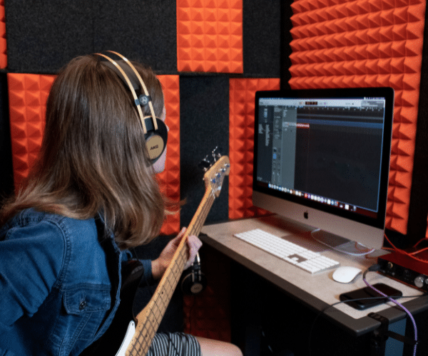 A student is shown recording bass inside a WhisperRoom at GCU's Recording Lab, engaged in the process of creating music.
