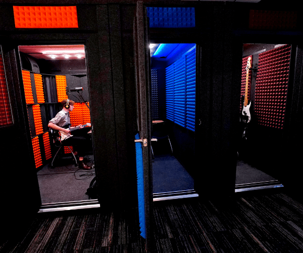 Three open WhisperRoom sound isolation booths featured inside GCU's Recording Lab, with one booth occupied by a guitarist wearing headphones, immersed in the recording process, demonstrating an active and dynamic creative environment.