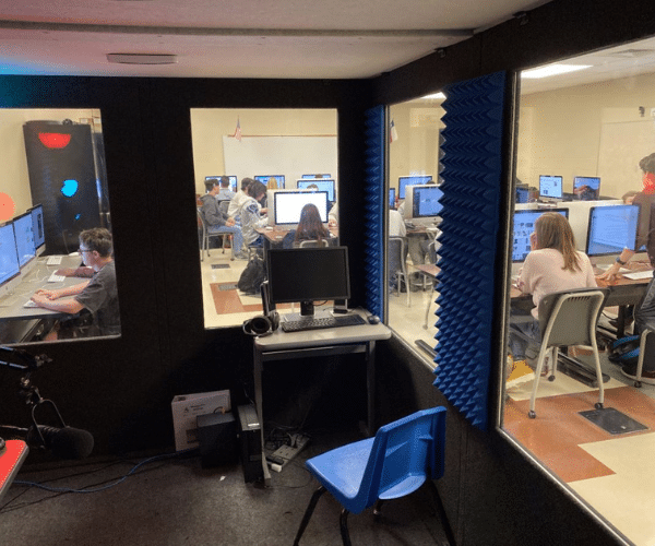 View from the interior of a WhisperRoom sound booth at Prosper High School, showing a classroom full of students each working at individual computers, emphasizing the interplay between creative isolation and collaborative learning.