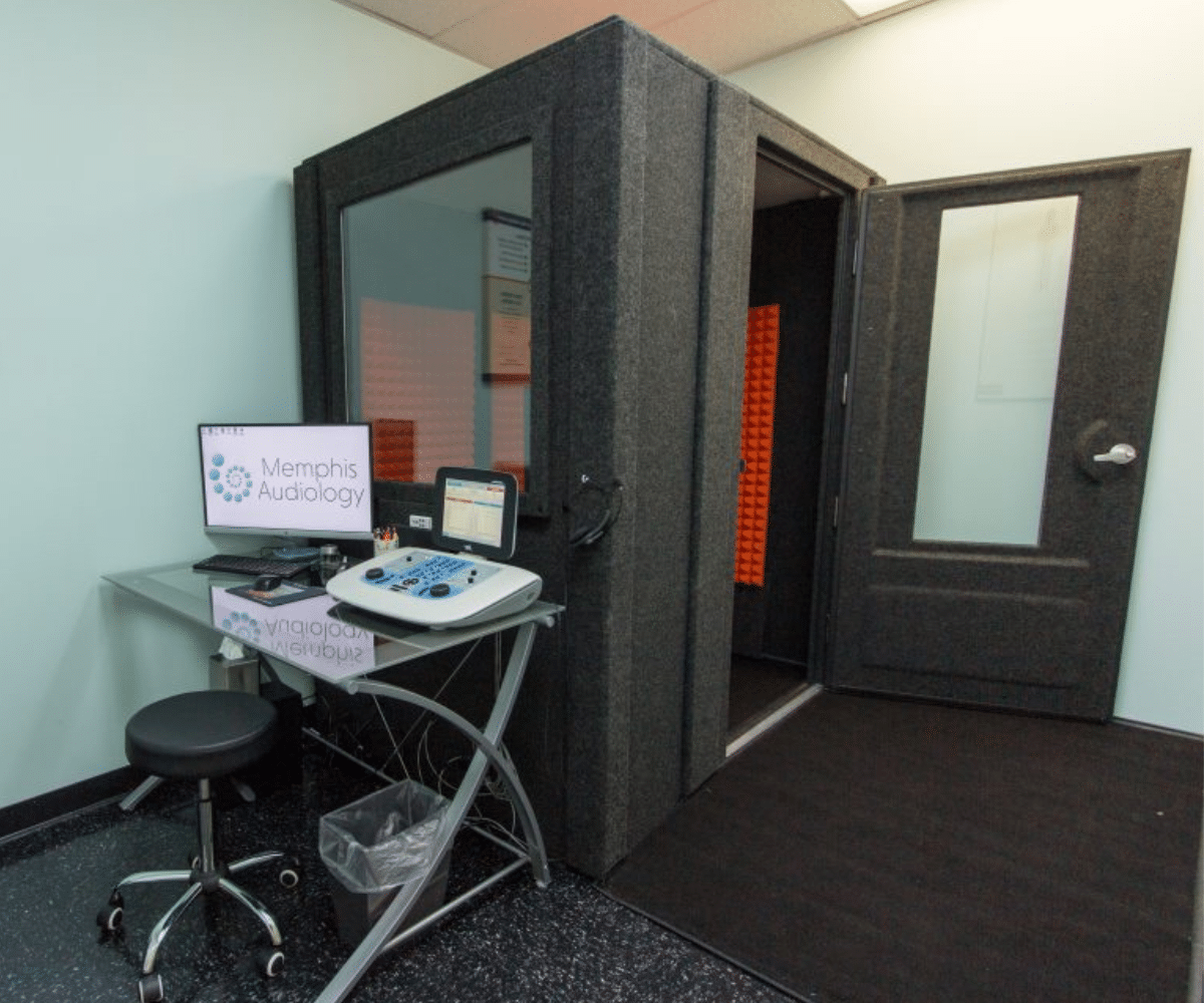 The WhisperRoom Audiology Basic Package displayed in Memphis Audiology's office. The audiometric booth is situated in the corner of the room, and the door is shown open. Adjacent to the booth's window, a small desk holds a computer and audiology testing equipment, creating a professional setup for precise evaluations and assessments.
