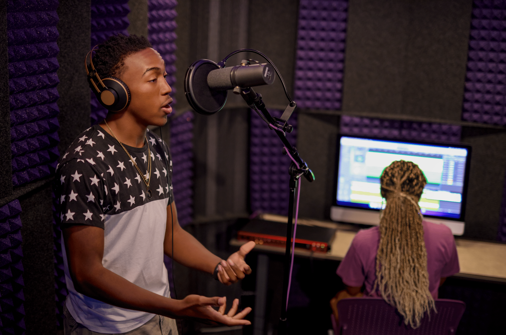 Two college students engaging in a recording session at GCU's Recording Lab. A young man is shown recording vocals into a condenser mic inside a WhisperRoom, while a female student operates the Digital Audio Workstation from a computer outside the booth