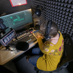 Featured image representing the 11 benefits of a WhisperRoom sound booth. The image showcases a college student inside a WhisperRoom booth at KEOM, recording a live radio broadcast. This visually illustrates the practical application of WhisperRoom booths in a professional radio broadcasting setting, highlighting the benefits of sound isolation, privacy, and optimal recording conditions for high-quality audio productions.