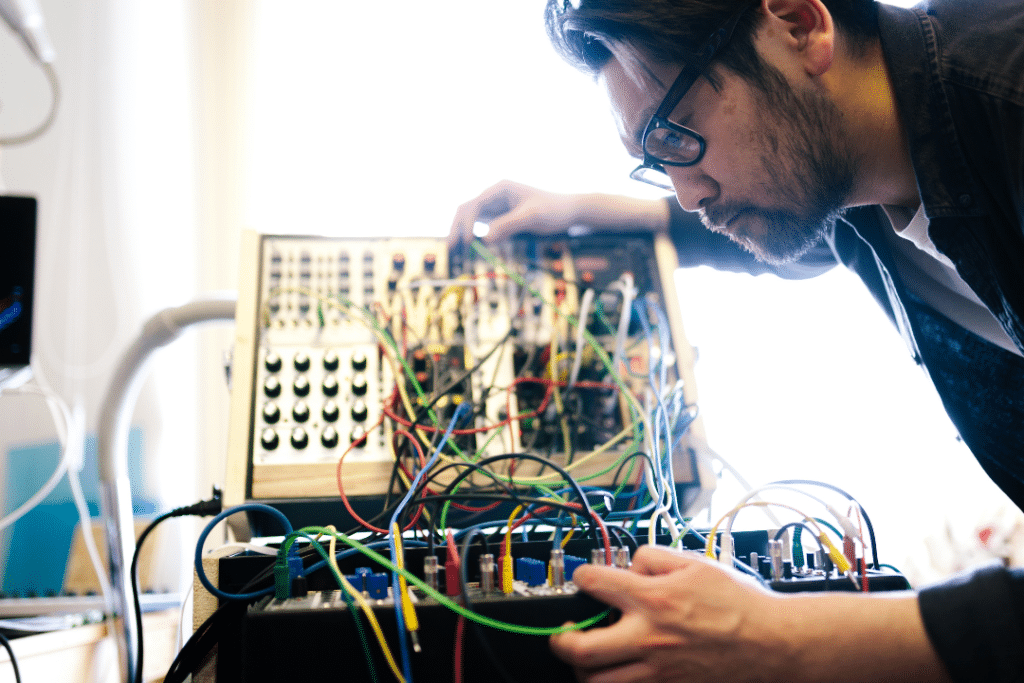 A man connecting analog synthesizers and audio equipment using patch cables. The image depicts the man's hands interconnecting various devices using colorful patch cables. The cables are being plugged into input and output jacks on the equipment, forming a complex network of connections. The image showcases the process of routing audio signals between different modules and components in a modular synthesizer setup, demonstrating the hands-on nature of analog audio patching.