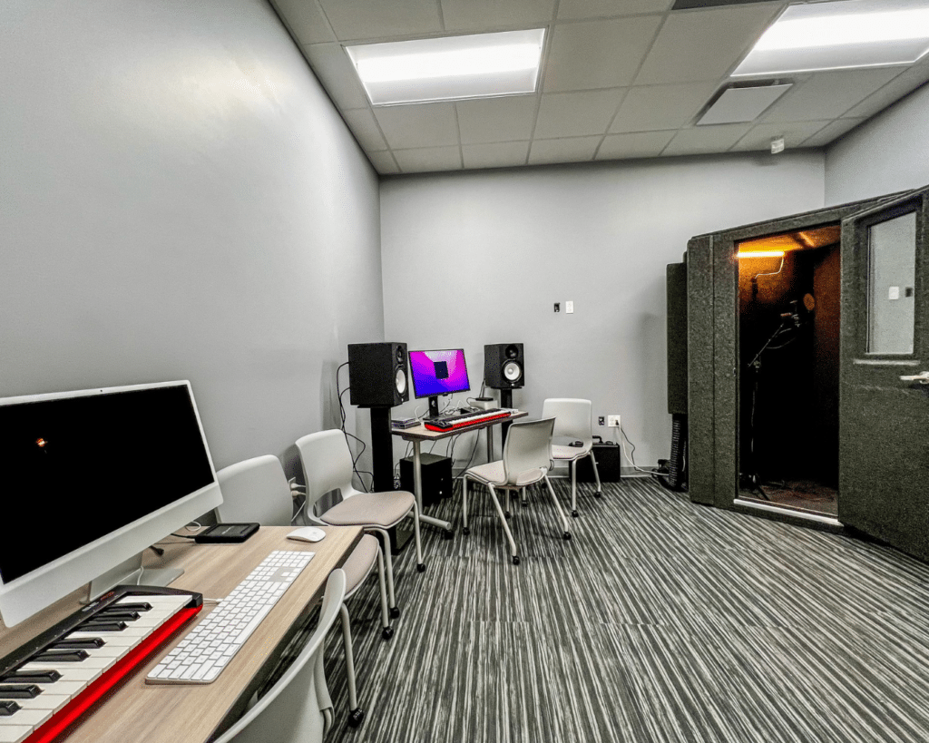 Image depicting a room within a music program setting, featuring a small WhisperRoom corner model located in the right-hand corner. Multiple music editing stations are visible along the walls outside the sound booth, showcasing a dynamic environment for music education and sound isolation.