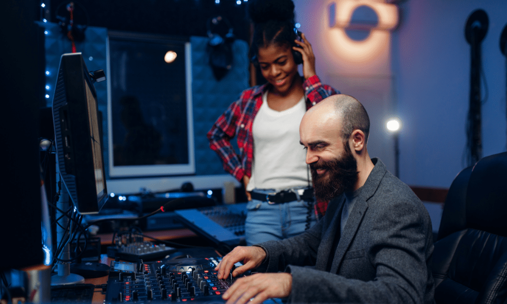 A music producer seated at a mixing board with a female musical artist standing next to him while listening to the mix, highlighting the importance of having a resilient space for recording.