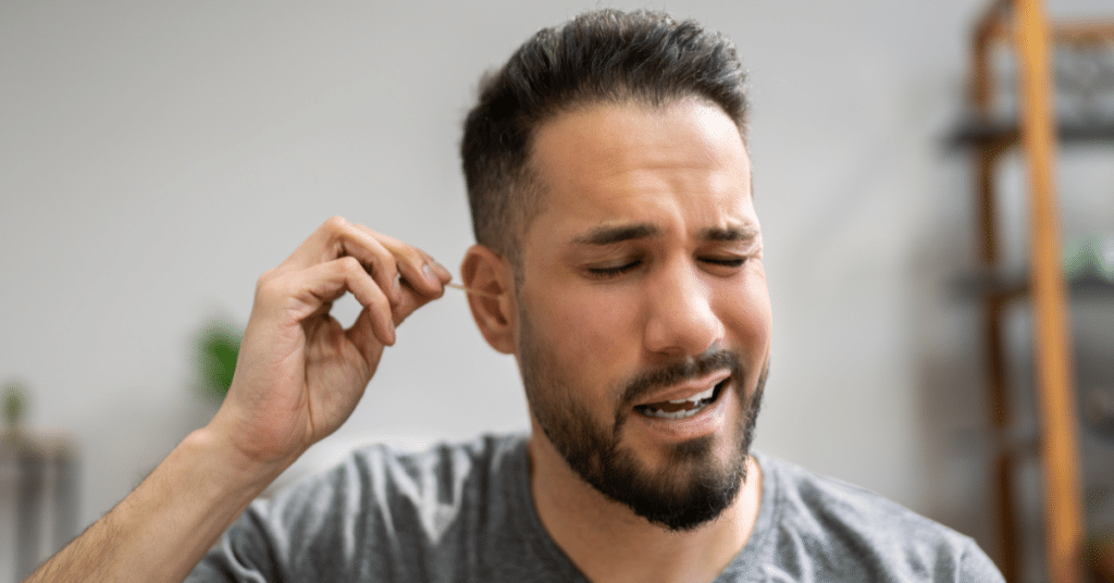 Image of man cleaning his ear with a cotton swab and making a wincing facial expression due to discomfort.